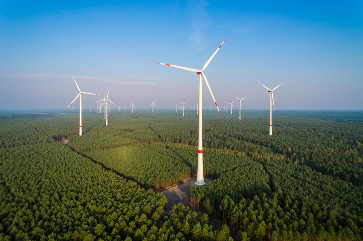 Windräder stehen im Grünen. Blauer Himmel über ihnen.