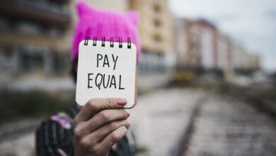 Nahaufnahme einer jungen Frau im Freien, die einen rosafarbenen Pussycat-Hut trägt und einen Notizblock vor sich hält, auf dem der Text pay equal geschrieben steht