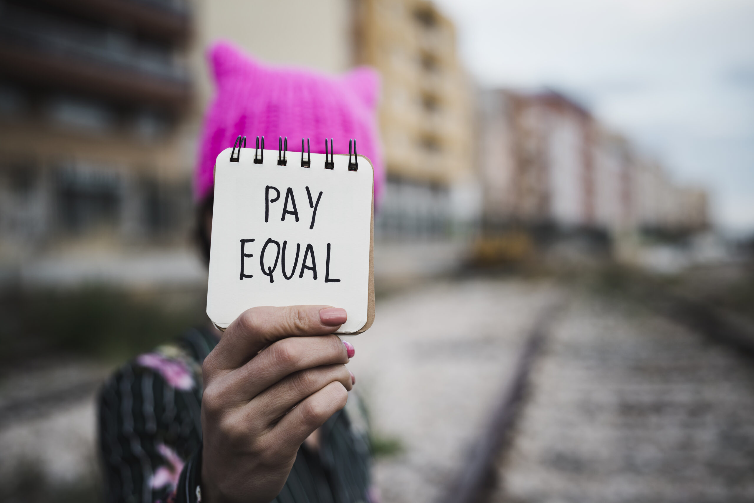 Nahaufnahme einer jungen Frau im Freien, die einen rosafarbenen Pussycat-Hut trägt und einen Notizblock vor sich hält, auf dem der Text pay equal geschrieben steht