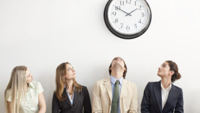 Vier Menschen in Bürokleidung sitzen und schauen zu einer Wanduhr.