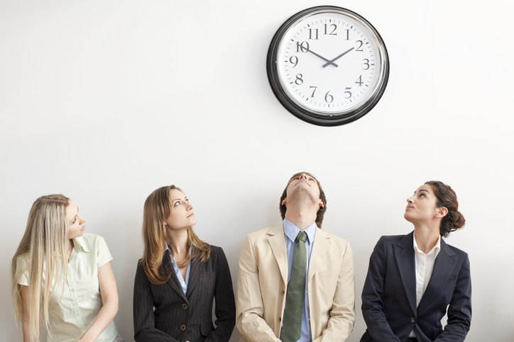 Vier Menschen in Bürokleidung sitzen und schauen zu einer Wanduhr.