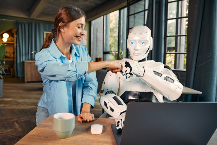 Darstellung Zusammenarbeit Mensch und KI: Frau gibt Roboter der am Laptop im Büro sitzt ein Fist Bump