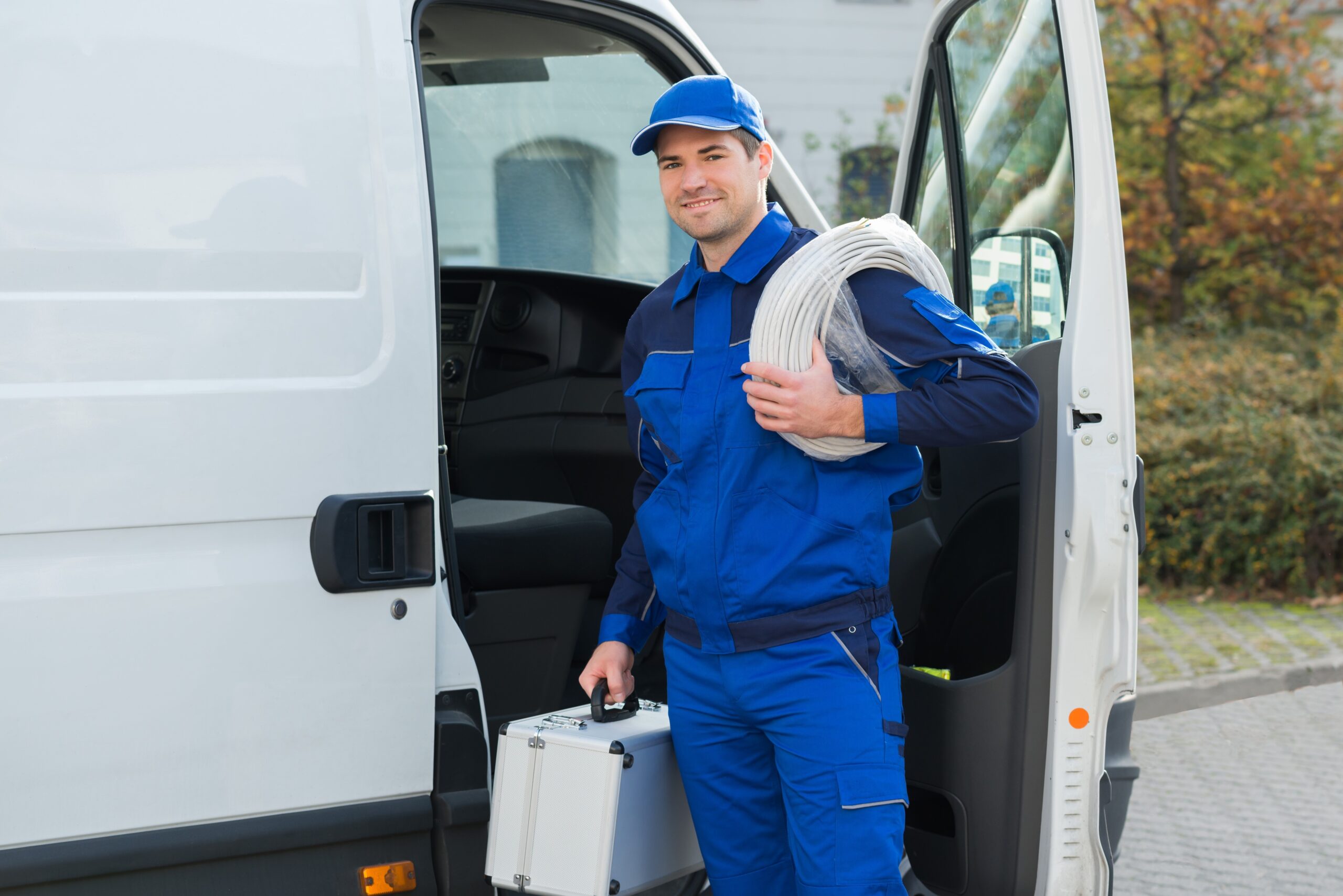 Ein Mann im Blaumann steht mit Werkzeugkoffer und Kabel vor seinem Auto.