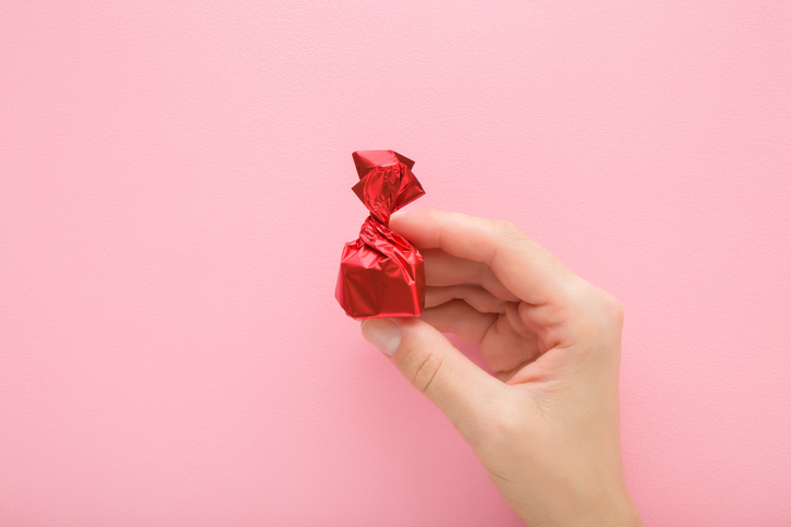 Junge erwachsene Frau Hand Finger halten und zeigen Süßigkeiten, die in leuchtend rotes Folienpapier auf hellrosa Tischhintergrund eingewickelt sind. Pastellfarbe.