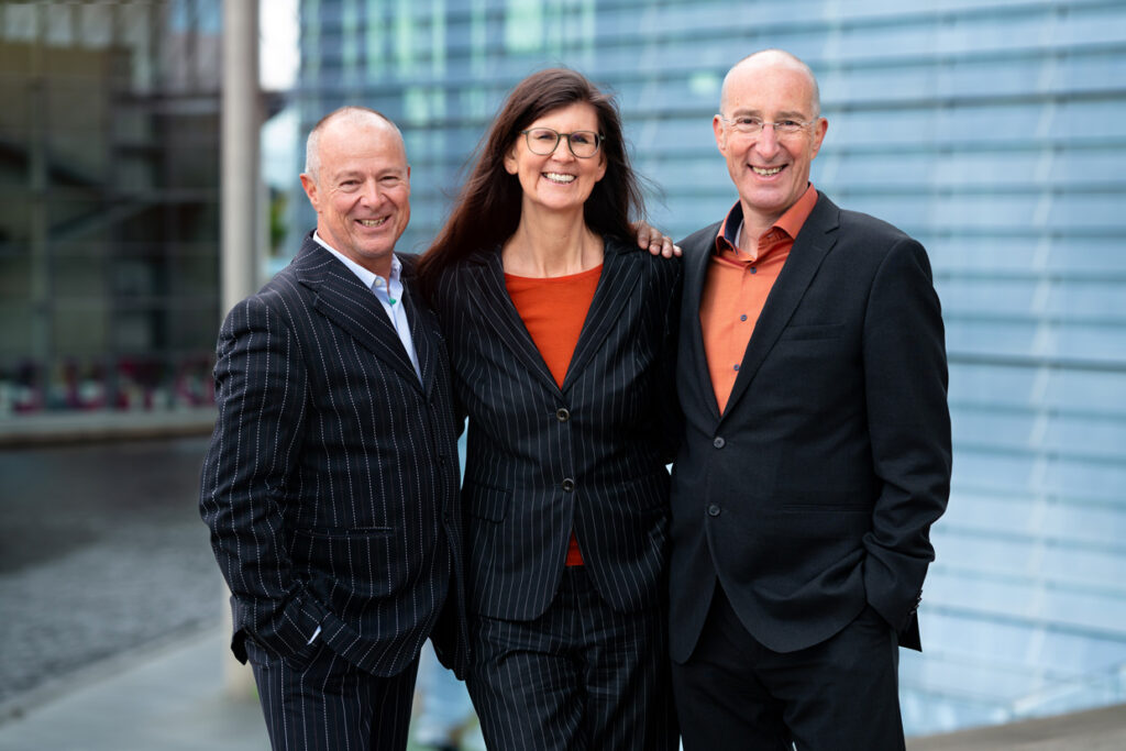 Drei Menschen in Anzug stehen lächelnd vor einem Gebäude.