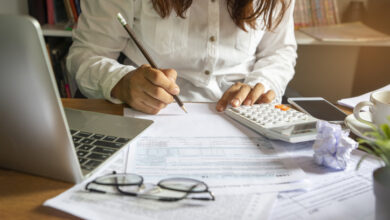 Buchhaltung bei der Budgetplanung: Aufnahme eines Schreibtisches mit Taschenrechner, Laptop und Brille sowie einer Person die dahinter sitzt und arbeitet