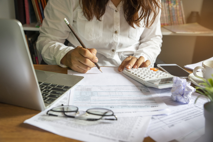 Buchhaltung bei der Budgetplanung: Aufnahme eines Schreibtisches mit Taschenrechner, Laptop und Brille sowie einer Person die dahinter sitzt und arbeitet