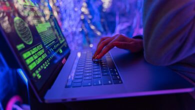 Cyberangriff dargestellt durch Hand auf Tastatur eines Laptops und grünem Code auf dem Bildschirm in einem blau-violett erleuchteten Zimmer