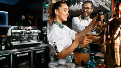 Young happy bartenders pouring beer from a beer tap while working in a pub. Focus is on woman.