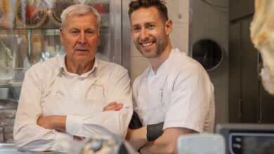 Two butchers are smiling with arms crossed in their butcher shop, with a meat display case behind them and various cuts of meat hanging