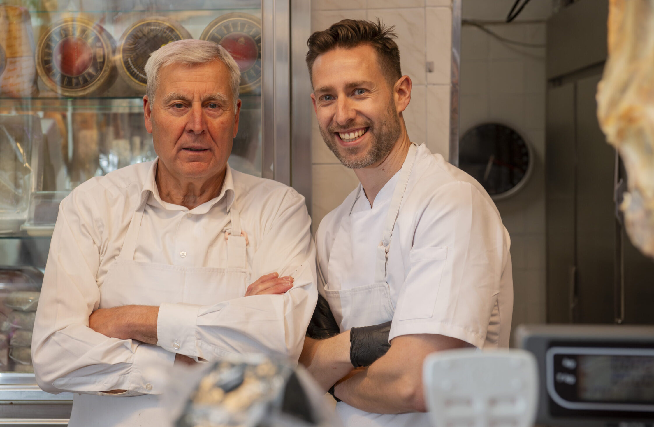 Two butchers are smiling with arms crossed in their butcher shop, with a meat display case behind them and various cuts of meat hanging